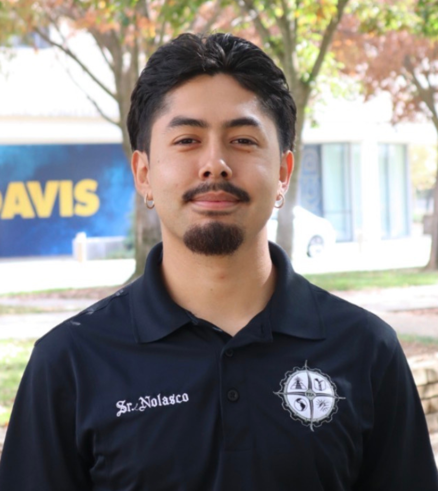 A man with dark hair and a goatee smiles, wearing a black shirt in an outdoor setting.